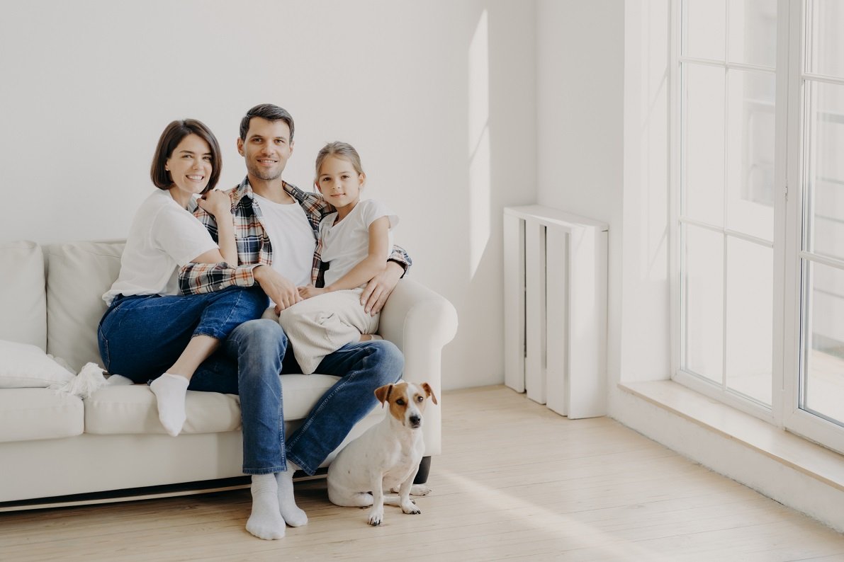 Family, togetherness and relationnship concept. Happy man embraces daughter and wife, sit on comfortable white sofa in empty room, their pet sits on floor, make family portrait for long memory